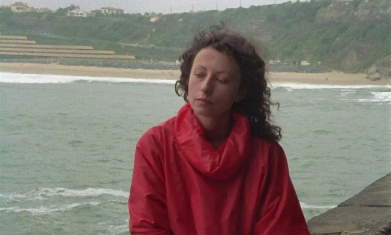 The Green Ray (1986) movie still: medium — A woman stands against a stone barrier near a sandy beach and ocean waves. She turns her head slightly…