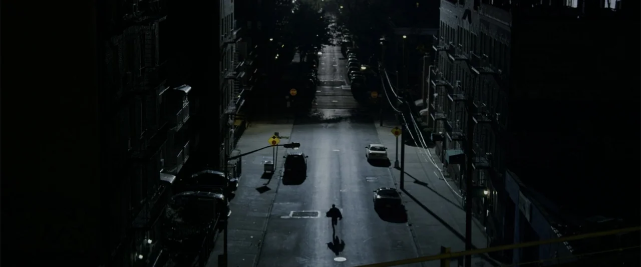The Amazing Spider-Man (2012) movie still: wide — An empty urban street at night with parked cars lines the tall dark buildings. A lone figure runs…