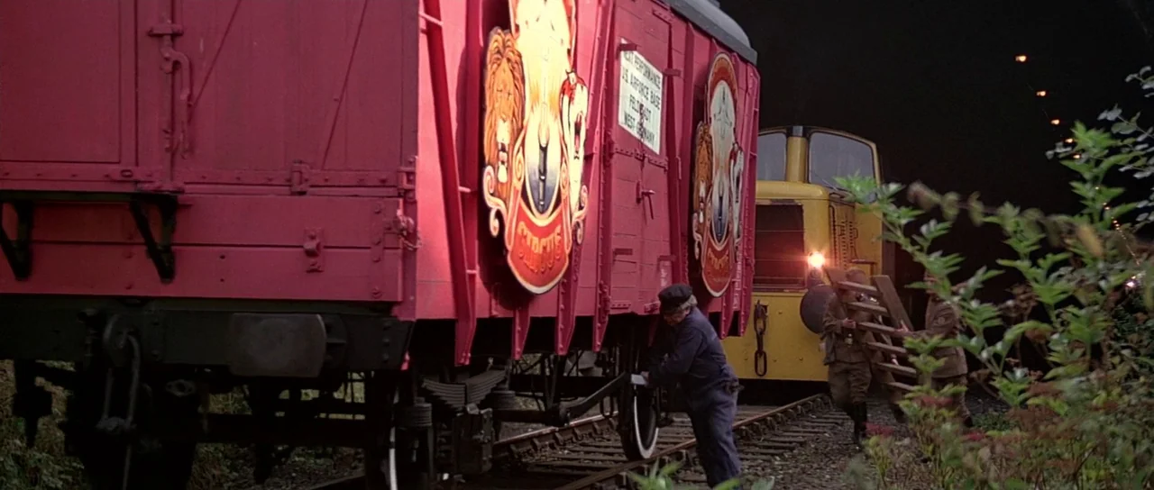 Octopussy (1983) movie still: wide — Bright red circus train cars stand on tracks leading into a dark tunnel. A worker inspects the wheel while two…