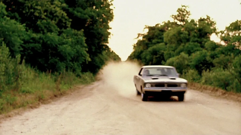 Undertow (2004) movie still: wide — A dusty unpaved road is flanked by dense green foliage under a warm sky. A vintage car drives towards the…