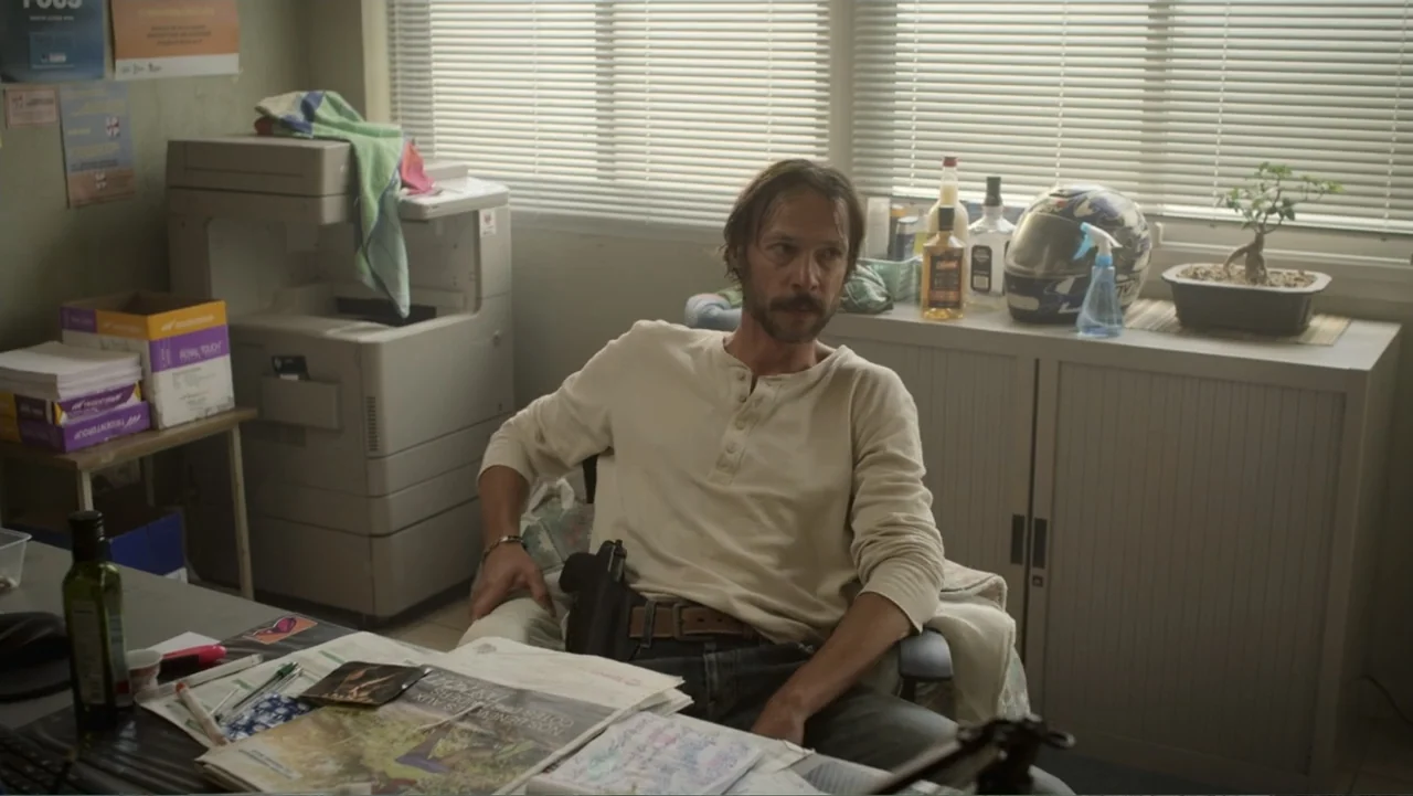 Unknown #1 movie still: medium — A cluttered small office space with a large photocopier, desk papers, and window blinds. A man sits in a chair at…