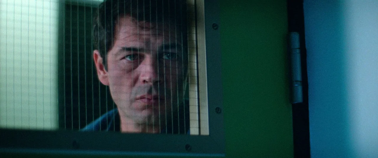Vigilante (1982) movie still: closeup — A man looks through a wire-reinforced glass partition into a plain, dimly lit room. The man's face is…
