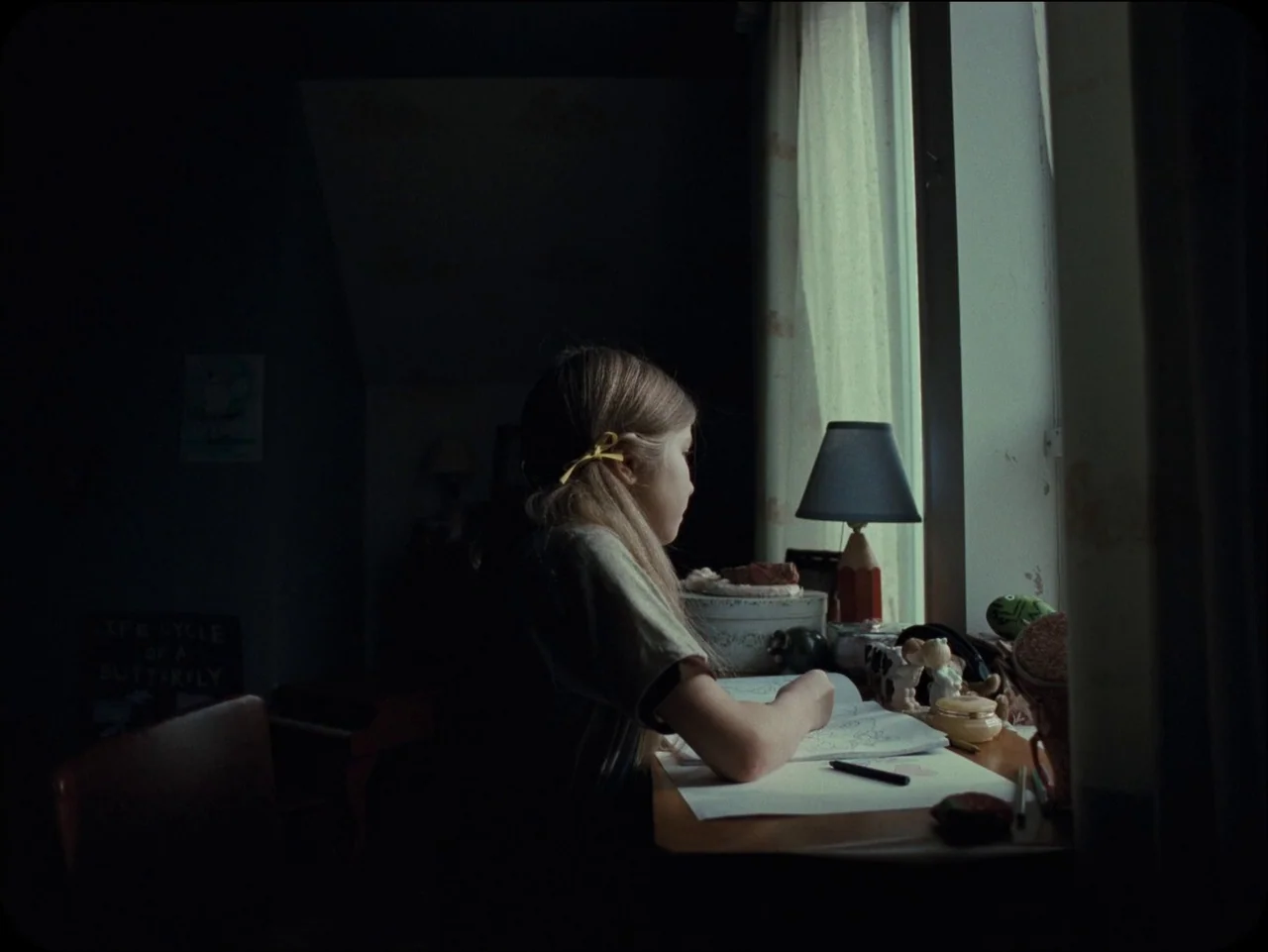 Longlegs (2024) movie still: medium — A dark, dimly lit room contains a small wooden desk positioned against a window. A young girl sits in profile…