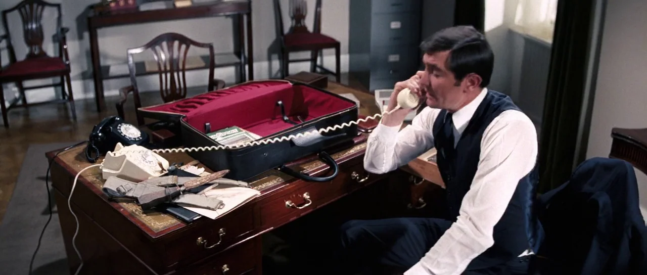 On Her Majesty’s Secret Service (1969) movie still: medium — A furnished professional office features wooden desks, period chairs, and a metal…