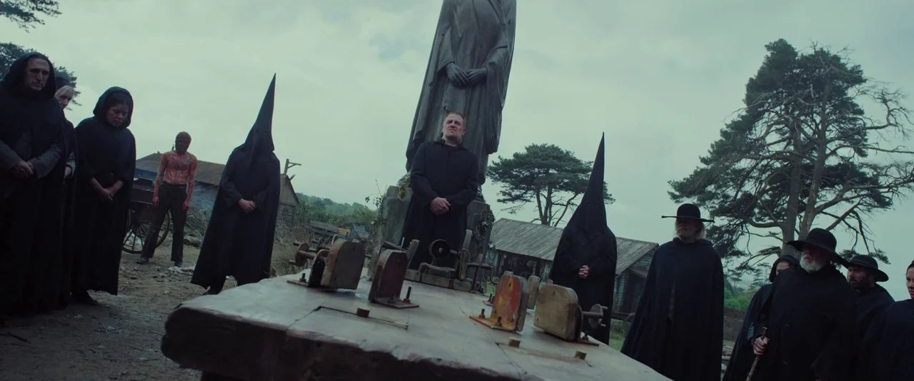 Apostle (2018) movie still: wide — A group of figures in dark robes gather in a muddy clearing before a statue. A man stands behind a wooden table…