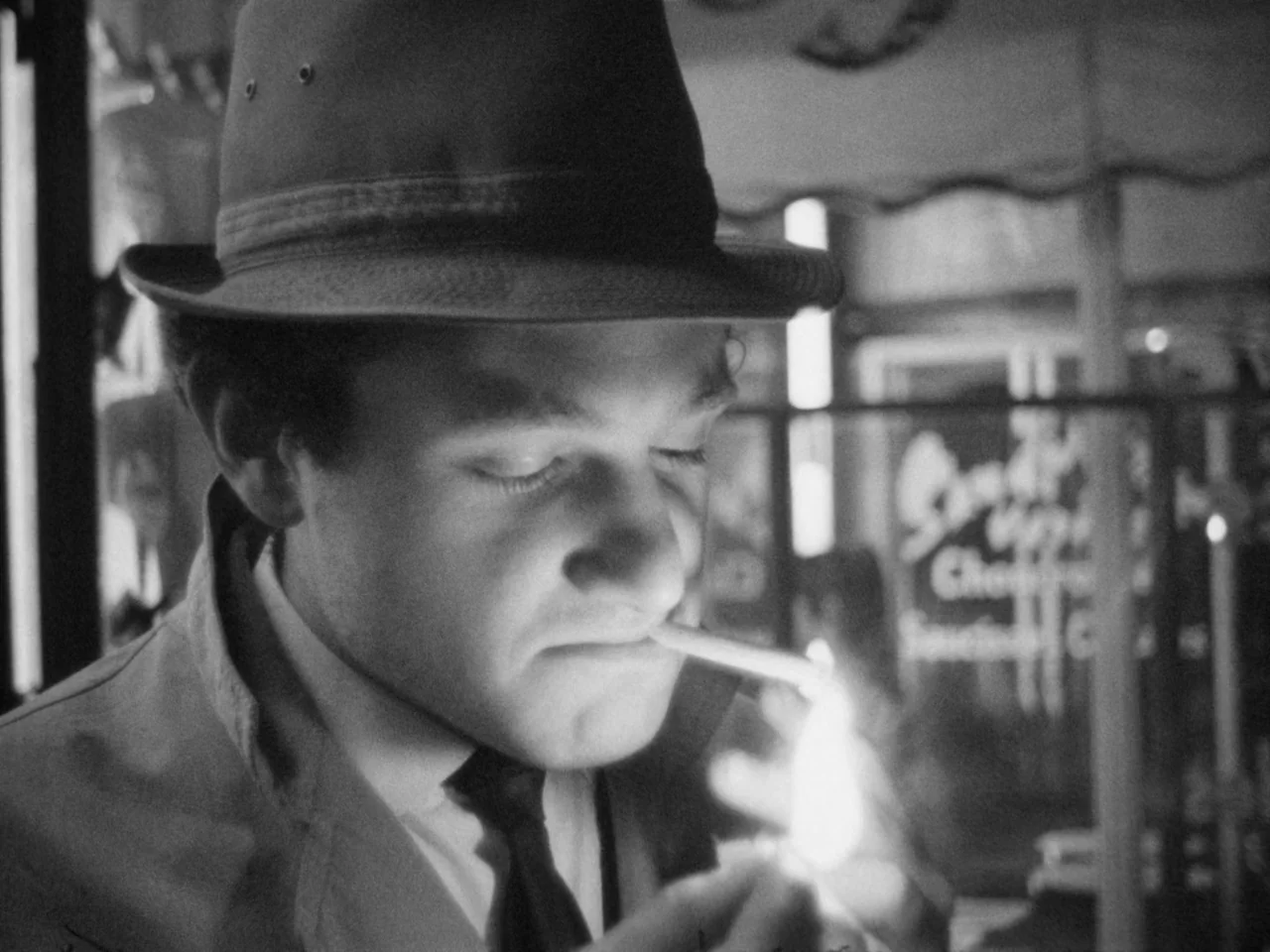 A Woman Kills (1968) movie still: closeup — A man in a fedora stands on a dark city street at night. He brings a lit lighter to the tip of his…