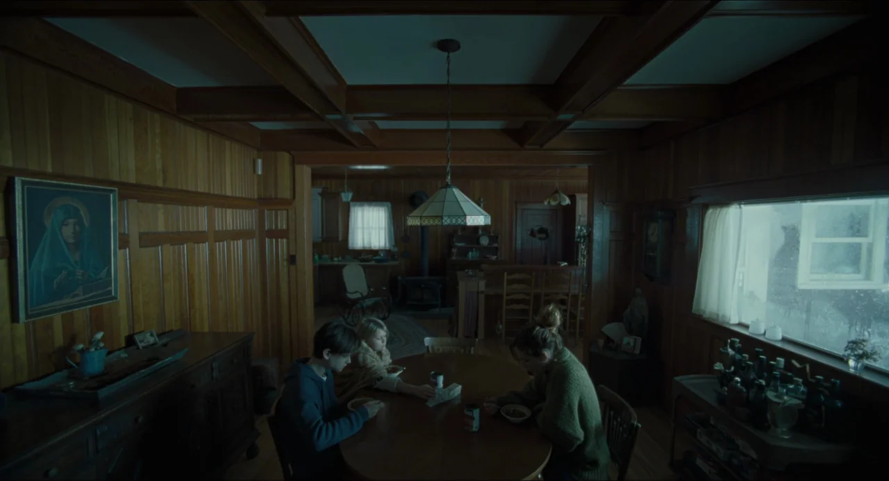The Lodge (2019) movie still: wide — A wood-paneled room features a round table surrounded by three seated people eating. A framed religious…