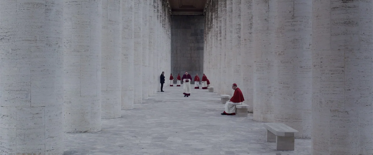 Conclave (2024) movie still: wide — A long symmetrical stone colonnade features multiple white cylindrical columns on either side. Several men in…