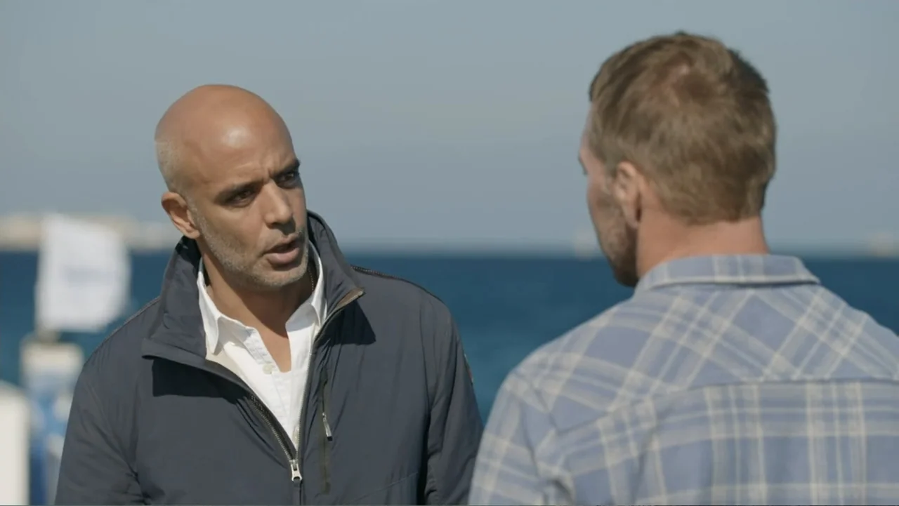 Serial Killer: Red Sea Attacks (2023) movie still: medium — A waterfront marina with a blue sea horizon visible in the background. One man speaks to…