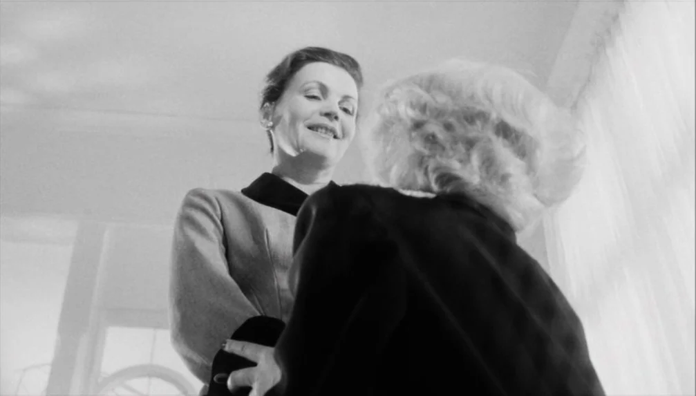 Veronika Voss (1982) movie still: medium — Two women interact in a bright, sparsely furnished interior with sheer white curtains. One woman in a…