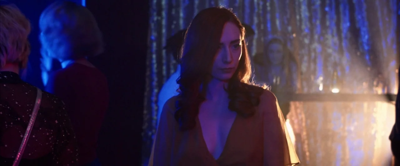 The Stylist (2020) movie still: medium — A dimly lit club with a metallic fringe curtain in the background. A woman stands in the foreground while…