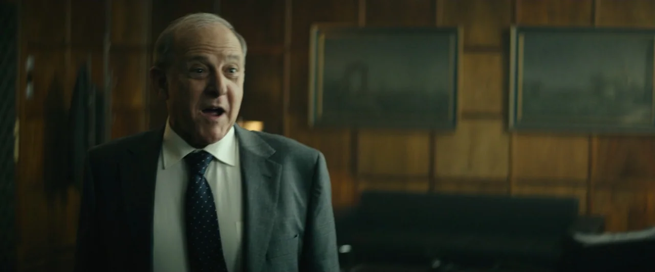 The Vault (2021) movie still: medium — An office interior features wood paneling and two framed landscape paintings on the wall. An elderly man in a…