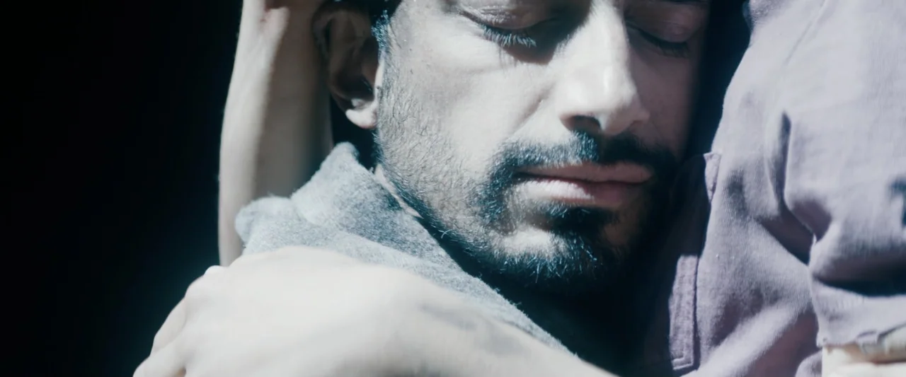 Encounter (2018) movie still: closeup — A dark, underexposed interior space features a close-up of two people embracing. The man with short dark…