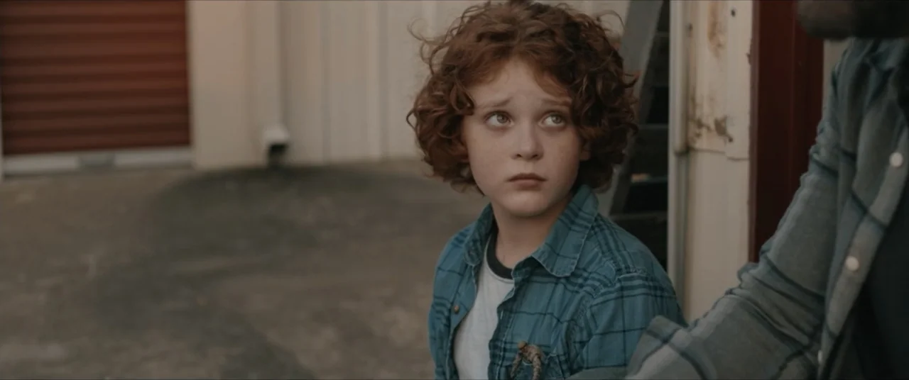 Possessions (2024) movie still: medium — A paved storage facility exterior features rows of metal roll-up doors under natural light. A curly-haired…