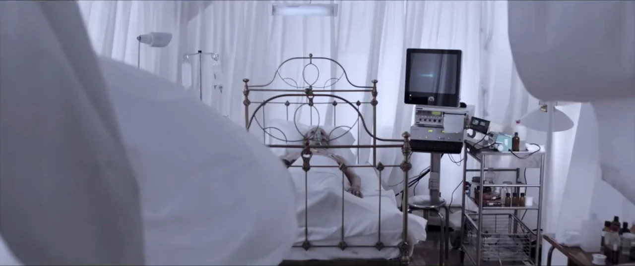 Room 6 (2006) movie still: medium — A clinical hospital room features a brass iron bed frame surrounded by white curtains. An elderly patient rests…