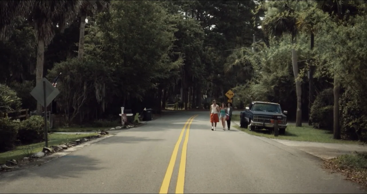 Eye for an Eye (2025) movie still: wide — A paved residential street is flanked by dense green trees and thick vegetation. Three teenagers walk side…