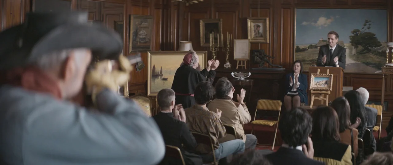 Daaaaaalí! (2023) movie still: wide — An upscale room features dark wood paneling, multiple framed paintings, and a podium. An auctioneer stands at…
