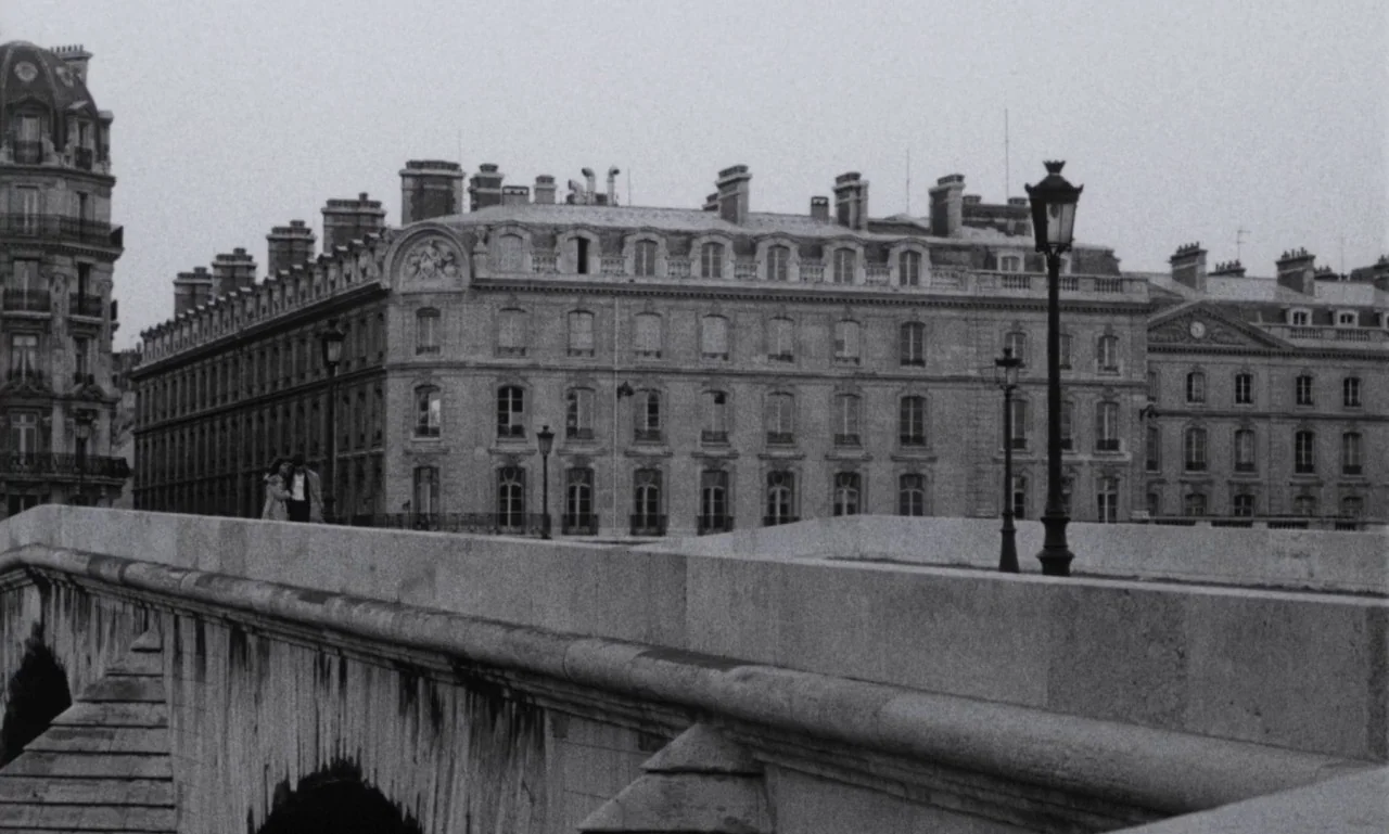The Secret Son (1979) movie still: establishing — A stone bridge extends across the frame toward multi-story Parisian apartment buildings. The sky…