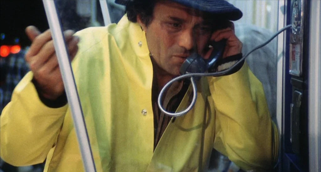 A Woman Under the Influence (1974) movie still: medium — A man stands inside a glass enclosure at night. He holds a black handset to his ear while…