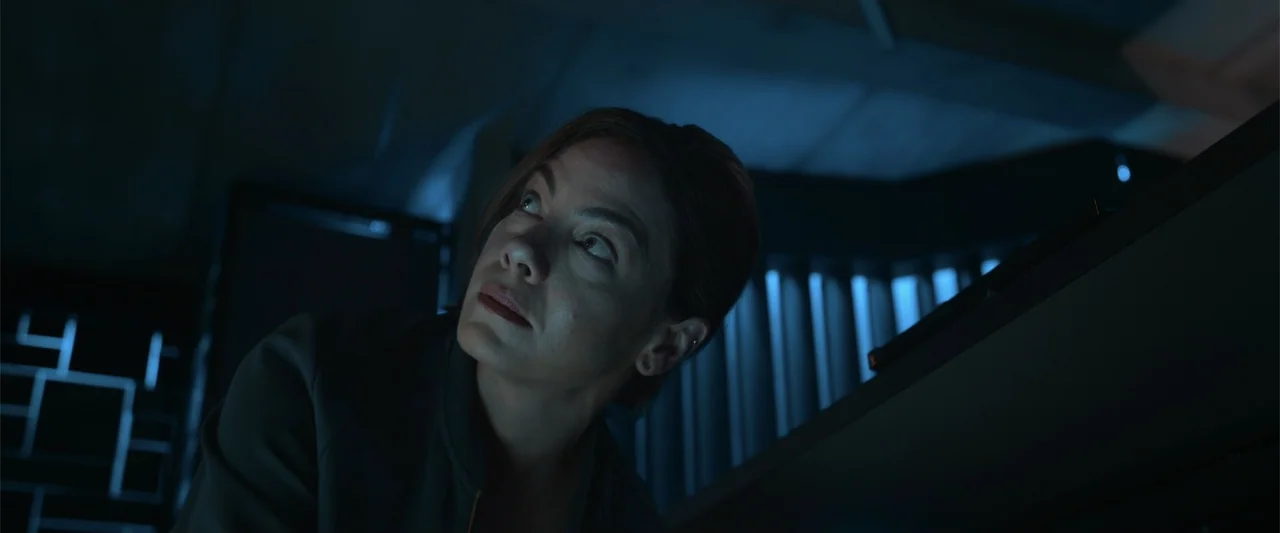 Black Site (2022) movie still: closeup — A dark interior space with rack-mounted server hardware illuminated by cool blue ambient light. A woman…