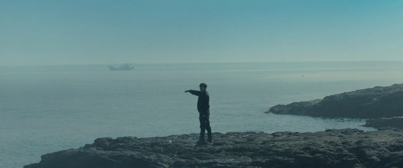 Decision To Leave (2022) movie still: wide — A man stands on a jagged rocky outcropping overlooking a calm, hazy ocean. A small distant fishing boat…