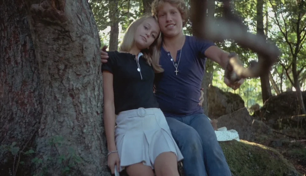 A Swedish Love Story (1970) movie still: medium — A young couple sits near the base of a large tree in a wooded area. The pair looks toward the…