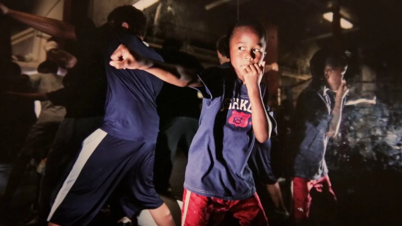 T-Rex (2015) movie still: medium — A dimly lit training area with multiple children practicing boxing movements in blue uniforms. The central boy…