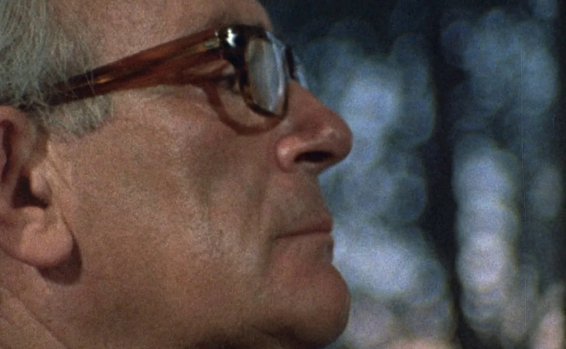 F for Fake (1973) movie still: closeup — A dimly lit room with an out-of-focus background of dark blue bokeh lights. An elderly man in glasses is…