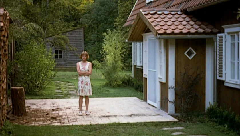 Funny Games (1997) movie still: fullbody — A rural backyard with a wooden house and a stone-tiled patio. A woman stands alone on the patio with her…