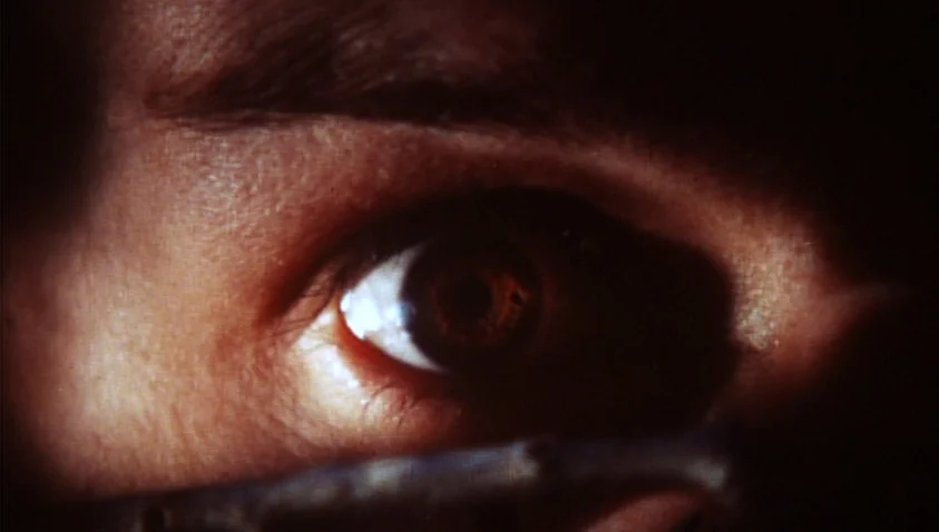 A Bay of Blood (1971) movie still: closeup — A close-up view of a single human eye framed in near-total darkness. The iris reflects a light source…
