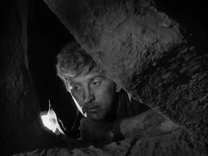 Ace In The Hole (1951) movie still: closeup — A rocky, dark interior space features a jagged aperture cut into solid stone. The man gazes forward…