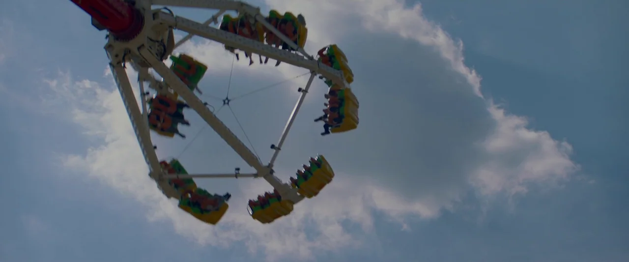 Sibyl (2019) movie still: wide — An amusement park ride rotates high against a bright blue sky with white clouds. The ride structure features…