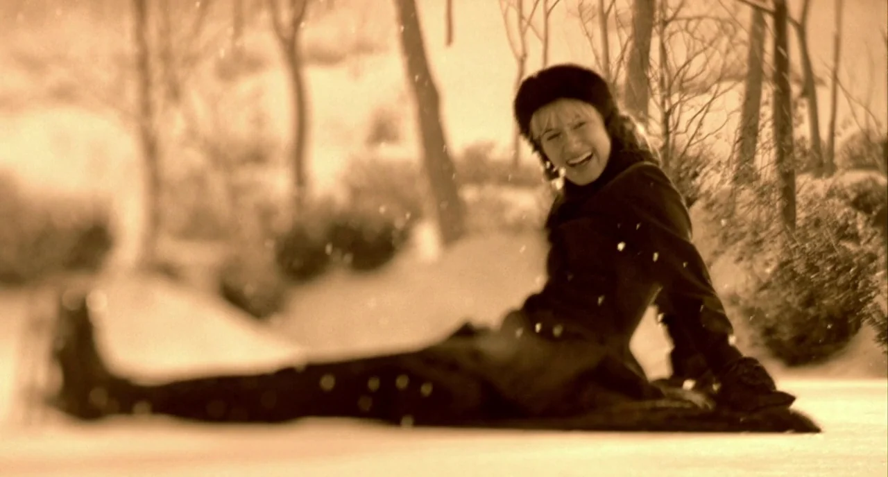 In The Cut (2003) movie still: medium — A woman sits on the snow-covered ground surrounded by bare trees in winter. She wears a dark coat and fur…