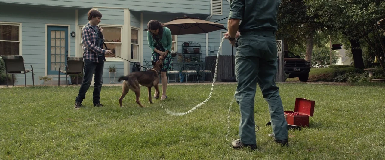 Max (2002) movie still: fullbody — A residential backyard sits behind a light blue house with a patio and lawn. A boy and a woman interact with a…