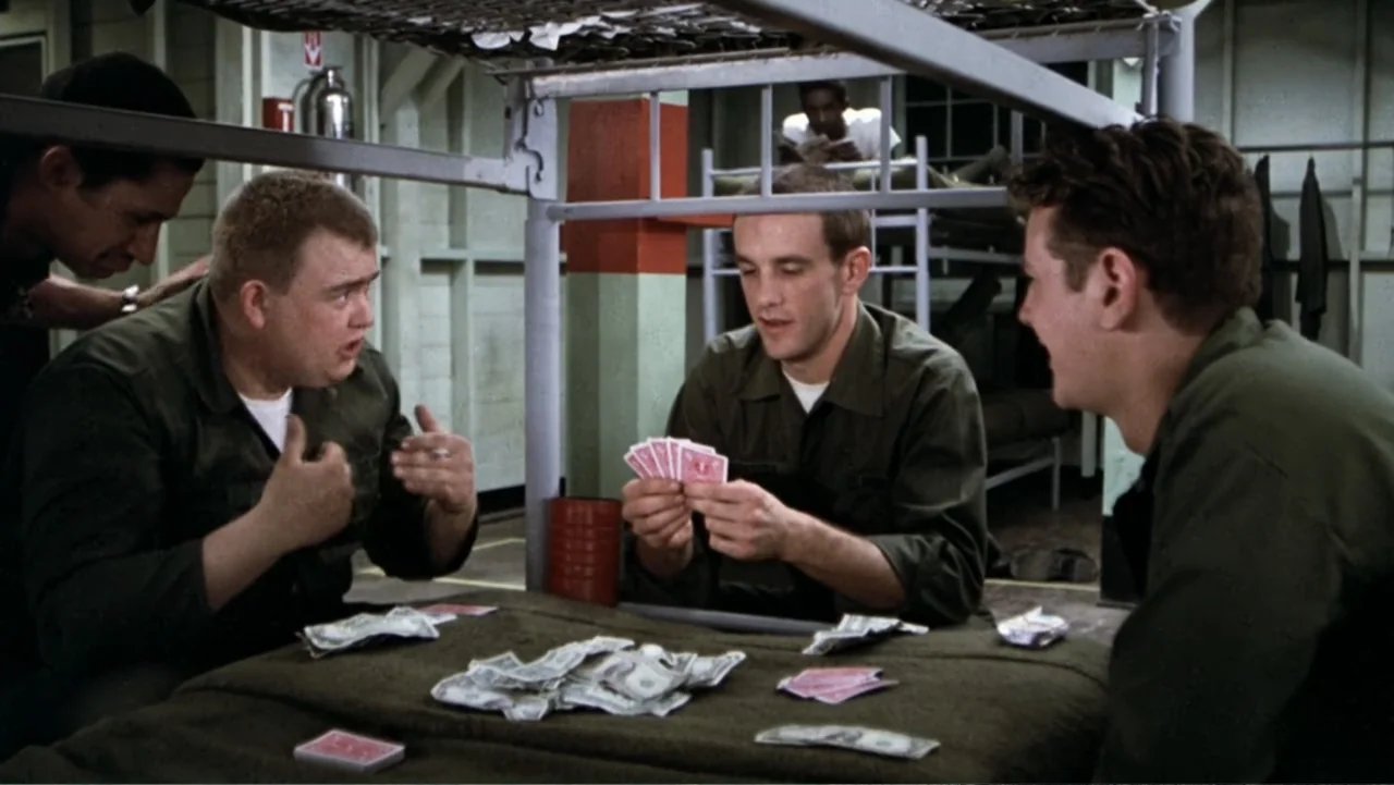 John Candy: I Like Me (2025) movie still: medium — A military barracks features bunk beds with simple frames and green mattress covers. Three…