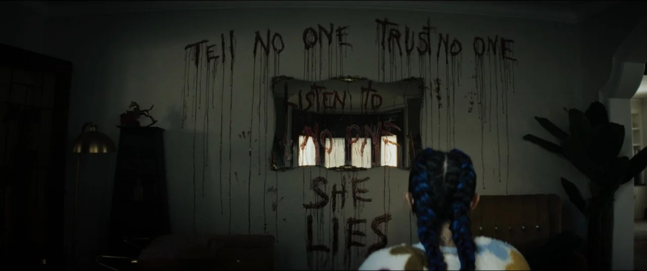 Vicious (2025) movie still: medium — A dark room features a wall covered in blood-like dripping text and a rectangular mirror. A person with blue…