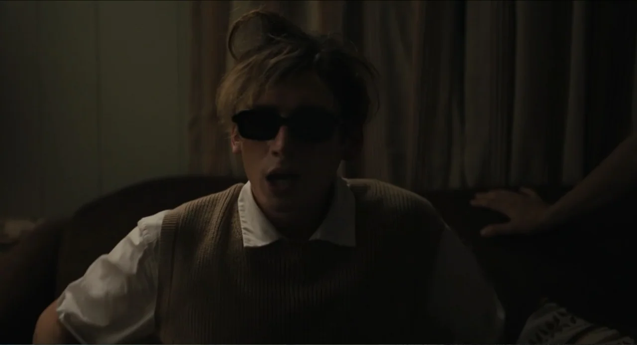 Hell of a Summer (2025) movie still: medium — A dimly lit indoor space features a dark sofa against gathered fabric curtains. A young man with messy…
