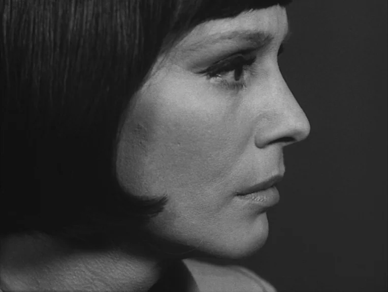 The Rite (1969) movie still: closeup — A dark, undifferentiated background sets the stage for a portrait-style headshot. A woman with a dark bob…