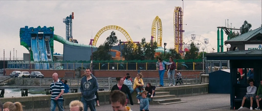 Happy-Go-Lucky (2008) movie still: wide — A seaside promenade features a public walkway in front of a colorful amusement park. Several people walk…