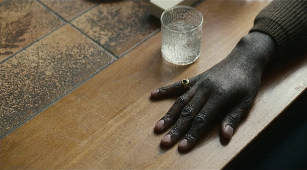 Red, White & Blue (2020) movie still: closeup — A wooden table surface sits next to tiled flooring in a domestic setting. A person's hand with a…