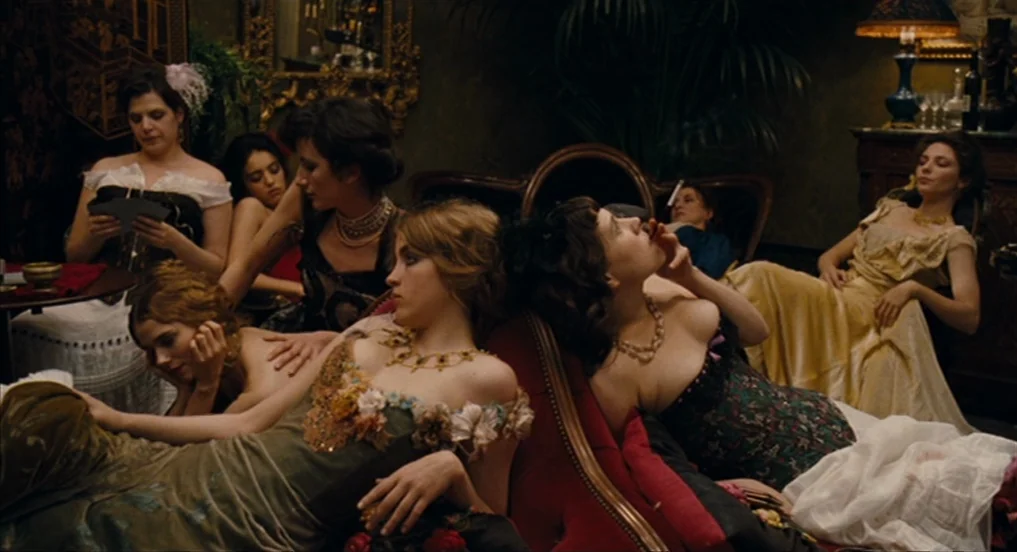 House of Tolerance (2011) movie still: medium — A dimly lit Victorian parlor features velvet couches, ornate gold-framed mirrors, and potted plants.…