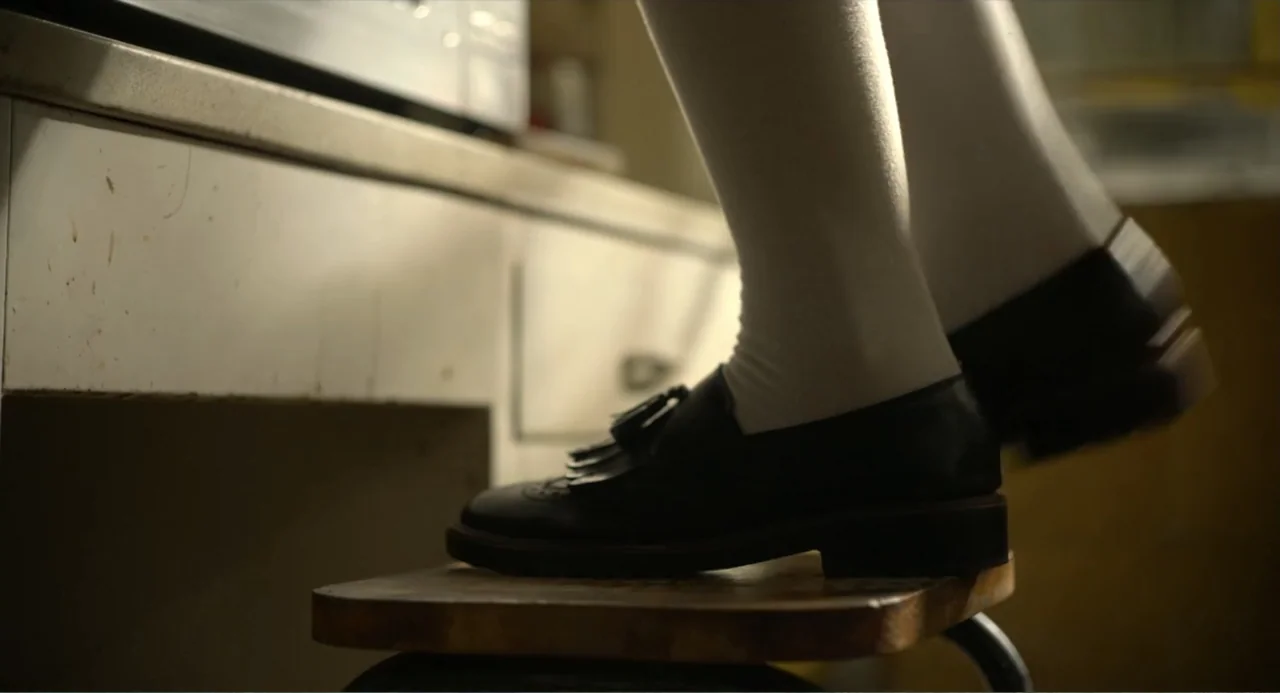 Heartbeat (2008) movie still: closeup — A close view of legs and feet standing on a wooden stool near a counter. White socks and black tassel…