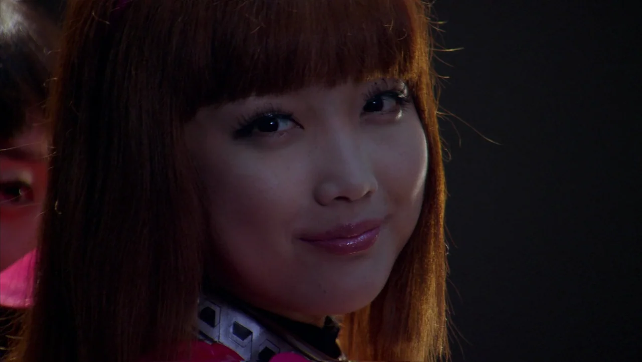 Cutie Honey (2004) movie still: closeup — A dark space features a young woman with reddish-brown hair and metallic armor. She looks directly at the…