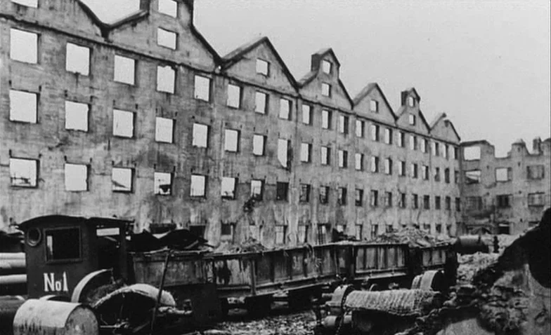 La Jetée (1962) movie still: establishing — A massive, multi-story brick factory stands gutted with empty window frames and roof damage. A small…