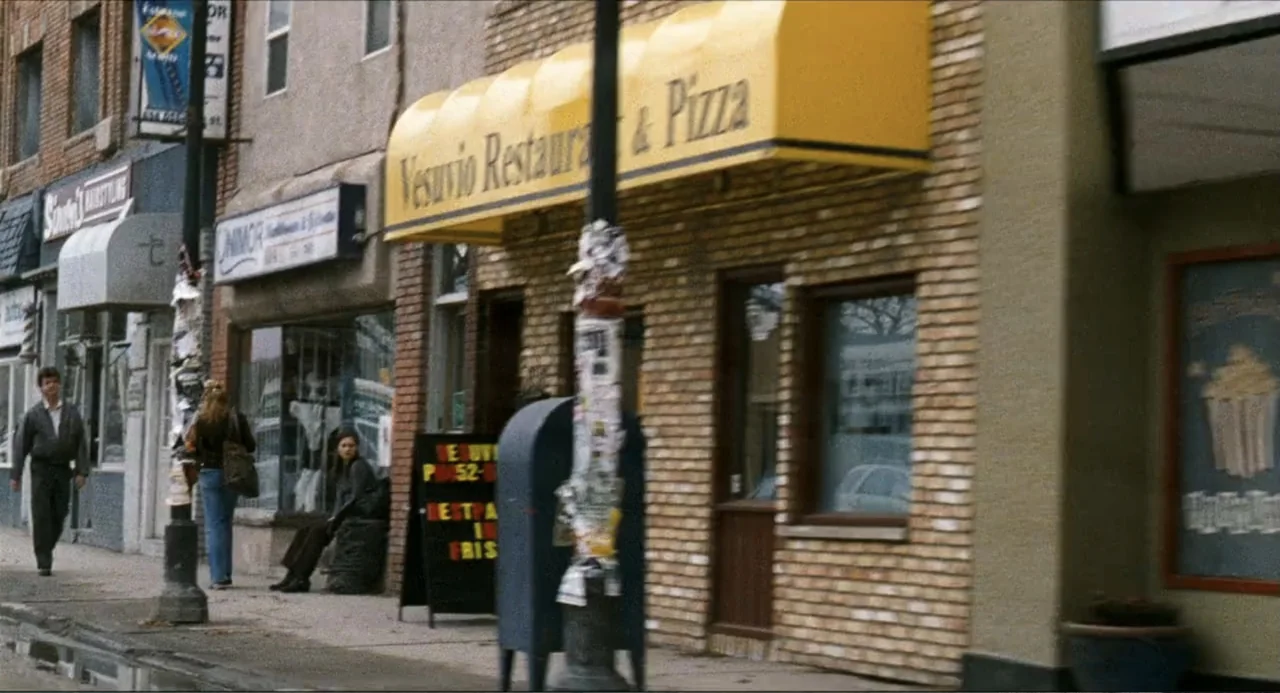 Colin Quinn: Red State, Blue State (2019) movie still: wide — A sidewalk fronting brick buildings features a yellow restaurant awning and store…