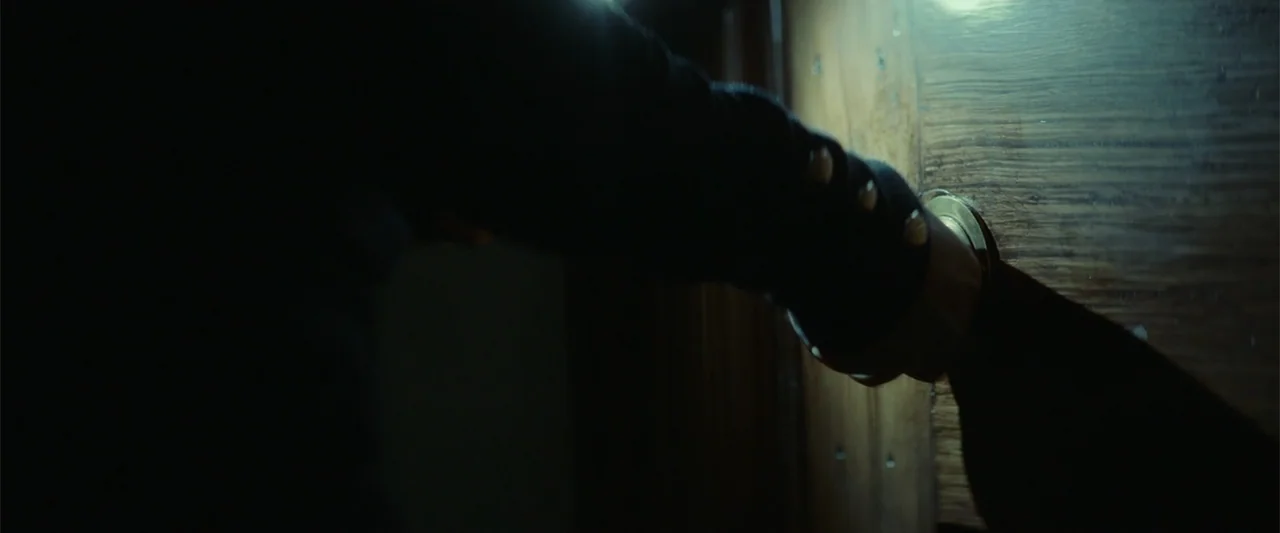 The Cellar (2022) movie still: closeup — A dimly lit interior view focuses on a hand grasping a brass door handle. The wooden door surface shows…