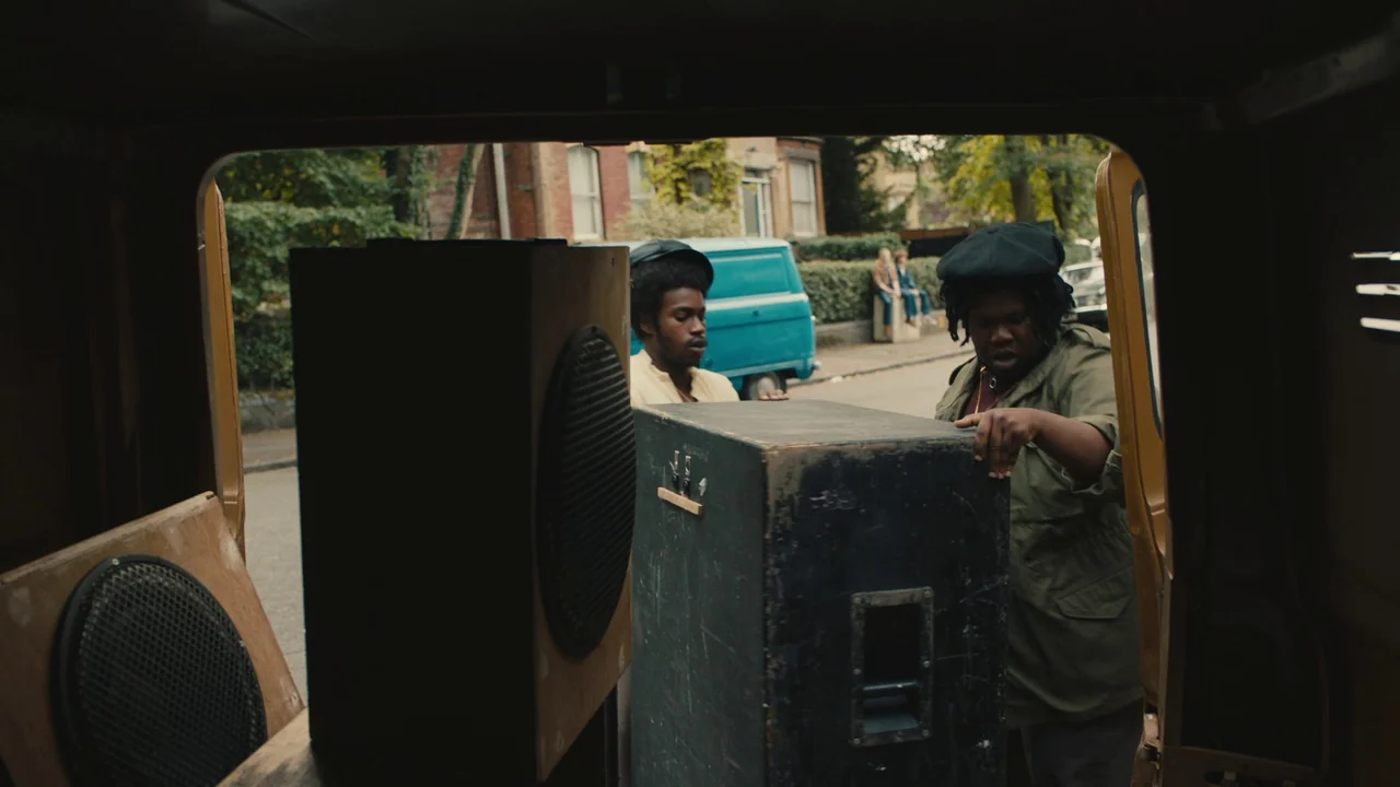 Lover’s Rock (2020) movie still: medium — A view from inside a van looking out at two men loading heavy speakers. The setting is a suburban street…
