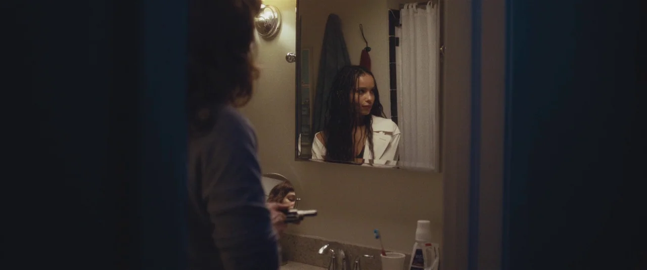 Gemini (2017) movie still: medium — A dimly lit room with muted beige walls and a rectangular mirror above the vanity. A woman with long dark braids…