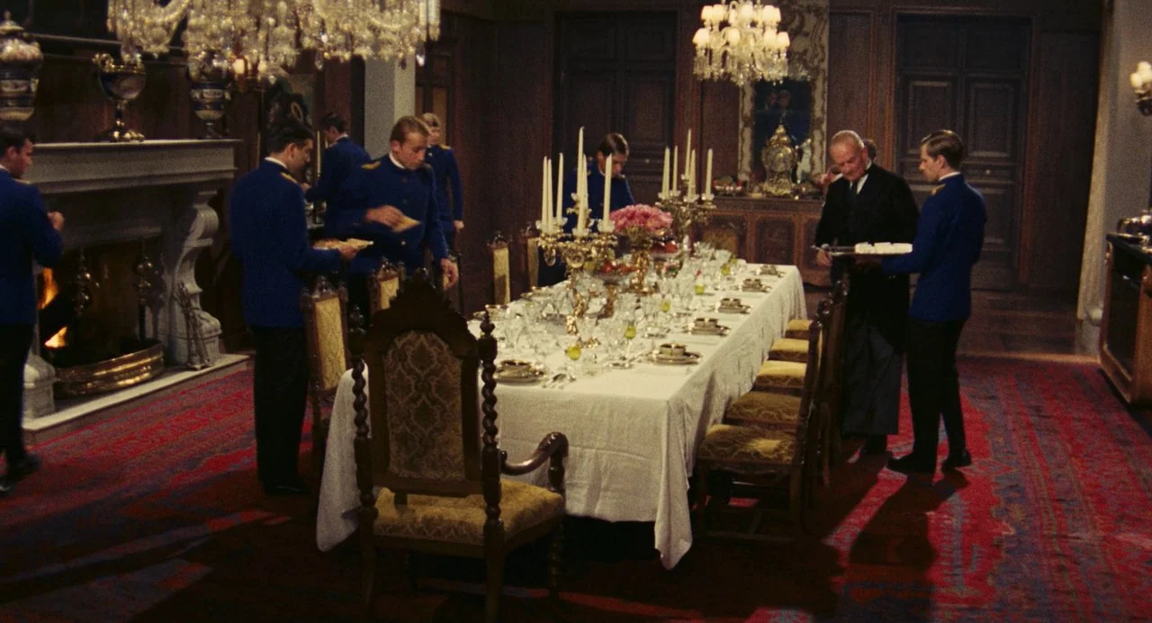 The Damned (1969) movie still: wide — An opulent dining room features a long table covered in white linen with crystal. Several men in blue formal…