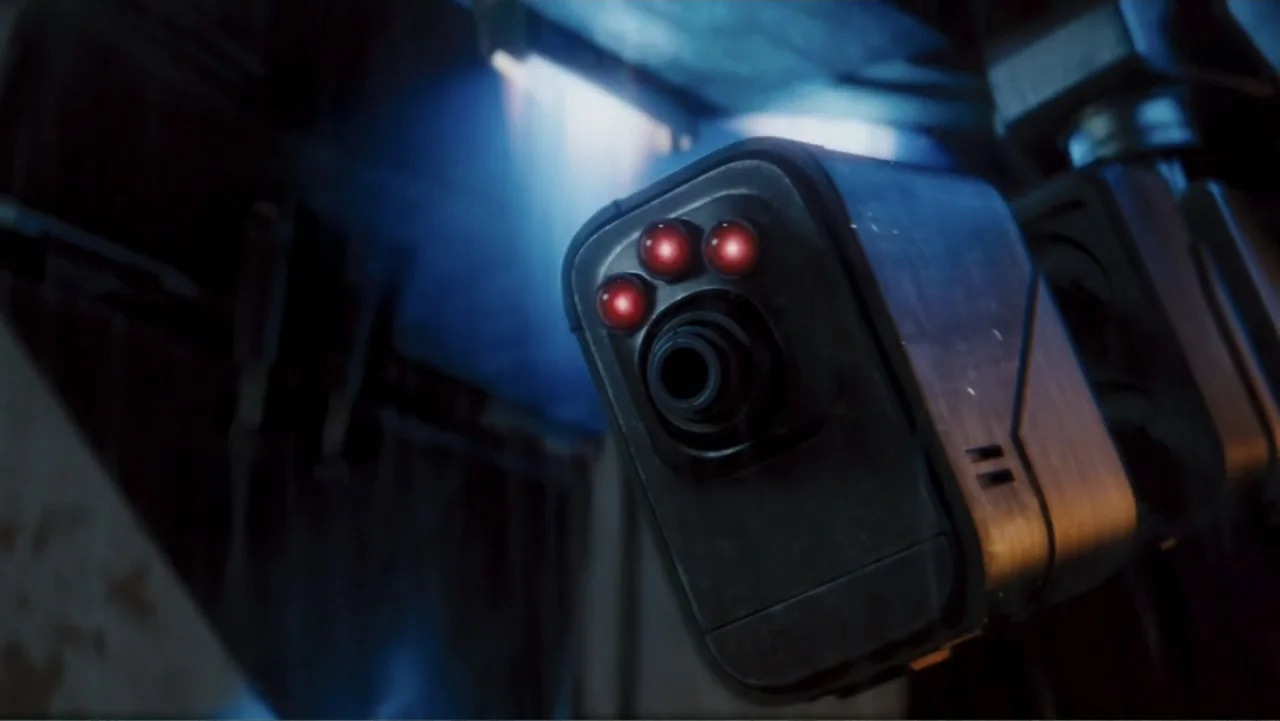 Robot Overlords (2015) movie still: closeup — A dark, industrial corridor contains a mounted electronic security device with glowing optics. The…